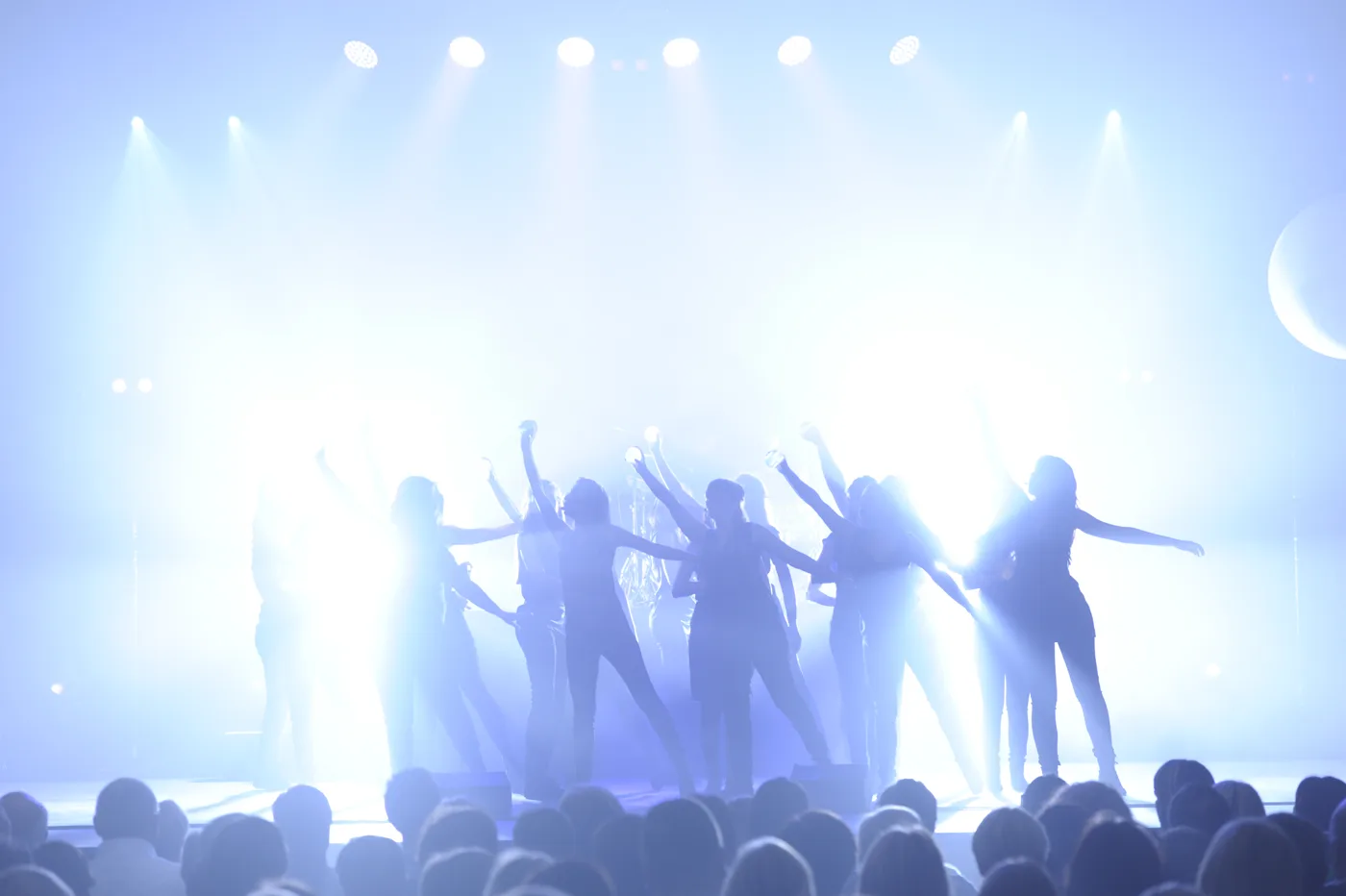 Singers with raised arms silhouetted against bright white stage lights, with audience heads in the foreground